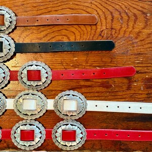 May include: A collection of leather belts with decorative silver-toned buckles. The belts come in various colors, including brown, black, red, and white. The buckles have a square center and a floral design around the edges.