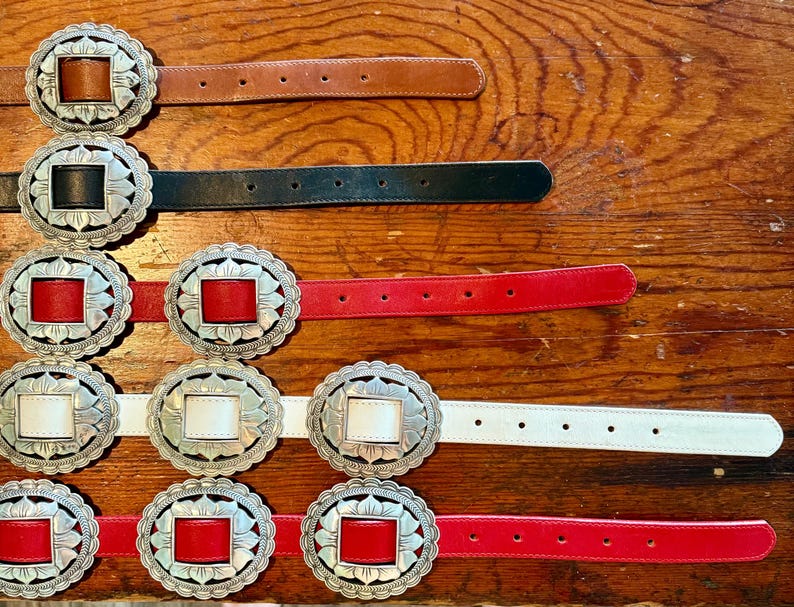 May include: A collection of leather belts with decorative silver-toned buckles. The belts come in various colors, including brown, black, red, and white. The buckles have a square center and a floral design around the edges.