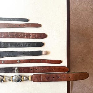 May include: An assortment of leather belts in various shades of brown and black, arranged on a white surface. The belts feature different textures, including embossed patterns and woven designs, with silver and gold-toned buckles.