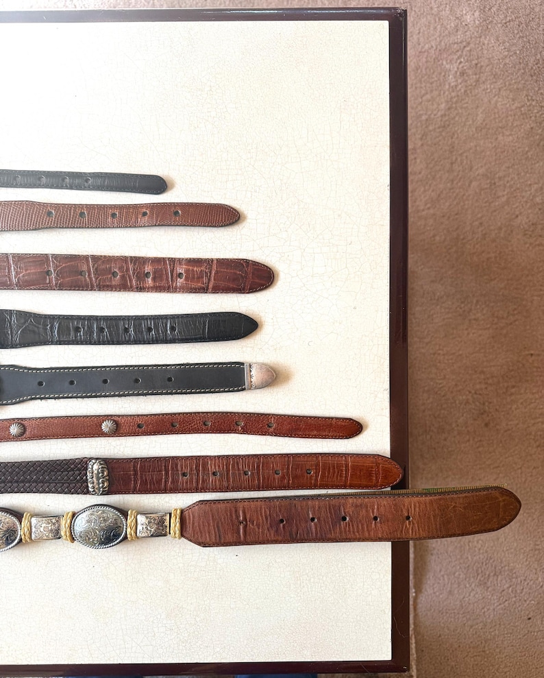 May include: An assortment of leather belts in various shades of brown and black, arranged on a white surface. The belts feature different textures, including embossed patterns and woven designs, with silver and gold-toned buckles.