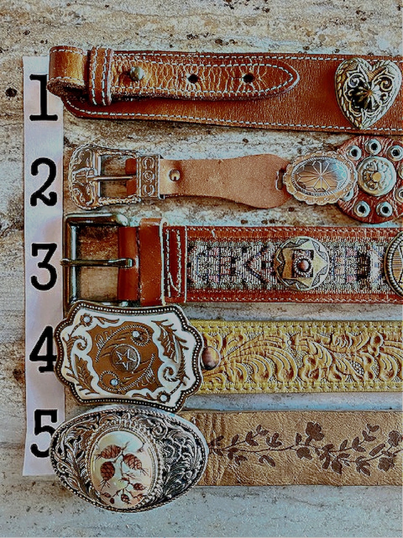 May include: An assortment of five leather belts with decorative buckles. The belts vary in color from light tan to brown, with intricate designs on the buckles, including floral patterns, stars, and heart shapes. The belts are arranged vertically.