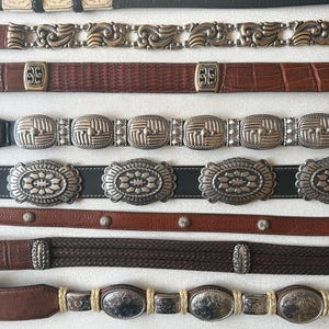 May include: A collection of vintage belts in various styles and materials. The belts include black leather with silver accents, brown leather with ornate metal buckles, and braided leather designs. The belts are displayed on a white surface.