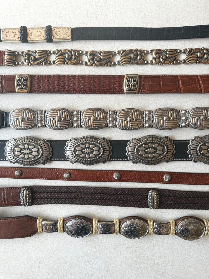 May include: A collection of vintage belts in various styles and materials. The belts include black leather with silver accents, brown leather with ornate metal buckles, and braided leather designs. The belts are displayed on a white surface.