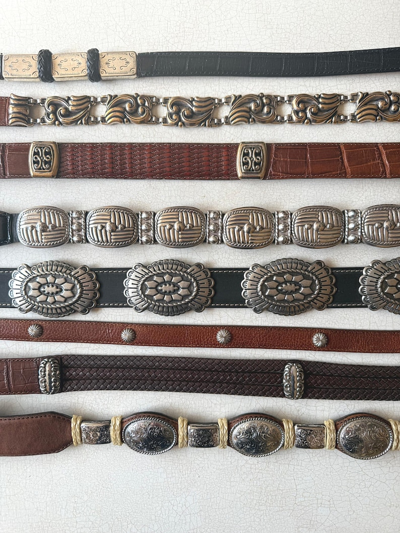 May include: A collection of vintage belts in various styles. The belts feature a range of materials, including leather and metal. Designs include ornate buckles, braided leather, and embossed patterns. Colors include black, brown, and silver.