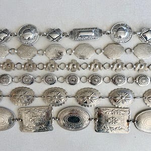 Puede incluir: Varios cinturones plateados con diversos diseños decorativos. Los cinturones presentan diferentes formas, incluyendo redondas, ovaladas y rectangulares, con patrones en relieve. Los cinturones están unidos por pequeños conectores circulares, creando un accesorio cohesivo.