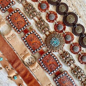 K&ouml;nnte beinhalten: F&uuml;nf verschiedene Lederg&uuml;rtel mit verzierten Metallschnallen und Verzierungen. Die G&uuml;rtel sind in einer Reihe angeordnet, wobei der oberste G&uuml;rtel ein braunes Lederband und eine gro&szlig;e, verzierte Metallschnalle hat. Die anderen G&uuml;rtel haben unterschiedliche Farben und Stile von Schnallen.