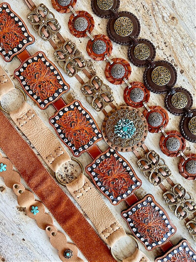 K&ouml;nnte beinhalten: F&uuml;nf verschiedene Lederg&uuml;rtel mit verzierten Metallschnallen und Verzierungen. Die G&uuml;rtel sind in einer Reihe angeordnet, wobei der oberste G&uuml;rtel ein braunes Lederband und eine gro&szlig;e, verzierte Metallschnalle hat. Die anderen G&uuml;rtel haben unterschiedliche Farben und Stile von Schnallen.