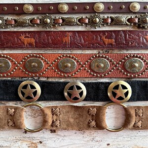Peut inclure: Cinq ceintures en cuir diff&eacute;rentes avec divers accents m&eacute;talliques. Les ceintures pr&eacute;sentent diff&eacute;rents motifs, notamment des &eacute;toiles, des cercles et des motifs animaliers. Les ceintures sont dispos&eacute;es en pile, la ceinture du haut &eacute;tant brune et la ceinture du bas &eacute;tant beige.