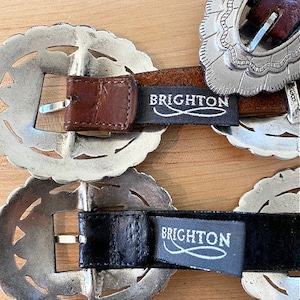 Może przedstawiać: Dwie srebrne klamry do paska z misternymi wzorami. Klamry są przymocowane do brązowych i czarnych sk&oacute;rzanych pask&oacute;w z nadrukiem "BRIGHTON" na sk&oacute;rze.