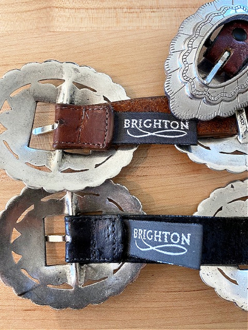 Może przedstawiać: Dwie srebrne klamry do paska z misternymi wzorami. Klamry są przymocowane do brązowych i czarnych sk&oacute;rzanych pask&oacute;w z nadrukiem "BRIGHTON" na sk&oacute;rze.