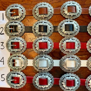 May include: Close-up of several belts with ornate silver buckles. The belts feature leather straps in brown, black, red, and white. The buckles have a floral design around a square or rectangular center.