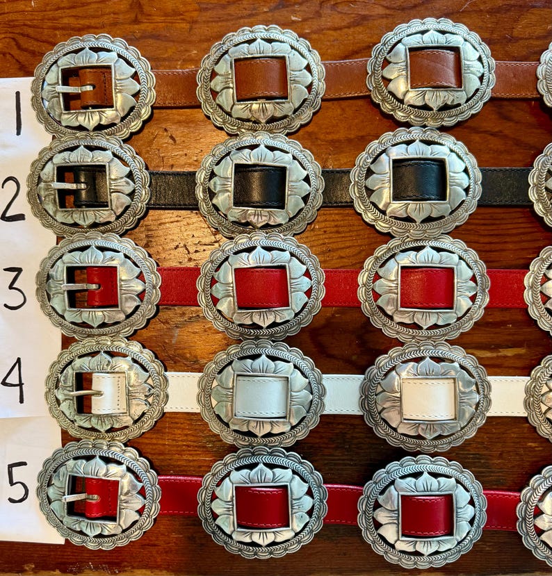 May include: Close-up of several belts with ornate silver buckles. The belts feature leather straps in brown, black, red, and white. The buckles have a floral design around a square or rectangular center.