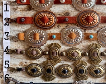 May include: A collection of Western-style belts with various buckle designs. The belts feature leather straps in shades of brown and black, paired with silver and gold-toned metal buckles. Some buckles have decorative details and colored accents.
