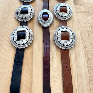 Może przedstawiać: Trzy sk&oacute;rzane paski z srebrnymi klamrami concho. Paski są czarne, brązowe i ciemnobrązowe. Klamry mają kwadratowy kształt z kwiatowym obramowaniem.