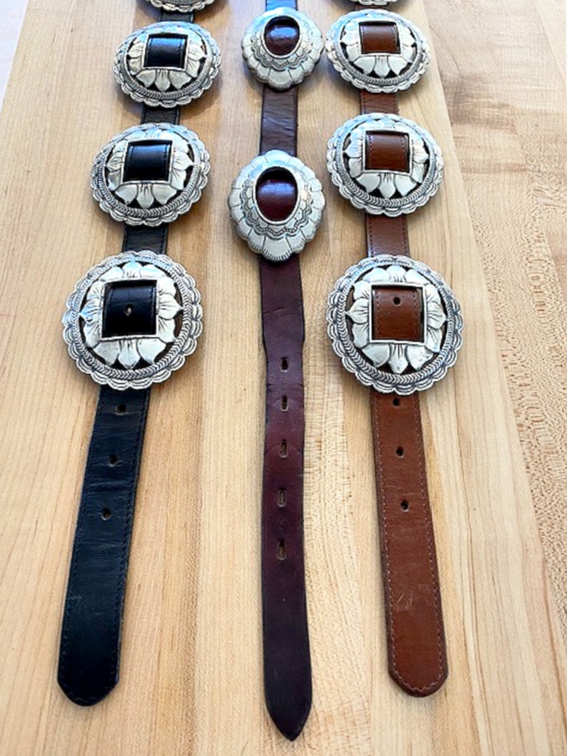 Może przedstawiać: Trzy sk&oacute;rzane paski z srebrnymi klamrami concho. Paski są czarne, brązowe i ciemnobrązowe. Klamry mają kwadratowy kształt z kwiatowym obramowaniem.