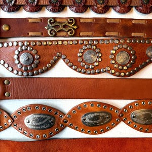 May include: Five brown leather belts with different decorative details. The top belt has round, embossed leather accents. The second belt has a metal buckle with a decorative design. The third belt has a scalloped edge with silver studs and round metal accents. The fourth belt has oval-shaped metal accents with a decorative design and silver studs. The bottom belt is plain.