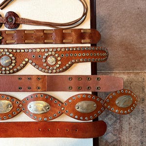 May include: Five brown leather belts with silver metal accents. The belts have different designs, including studs, circles, and oval shapes. The belts are arranged in a stack on a white surface.