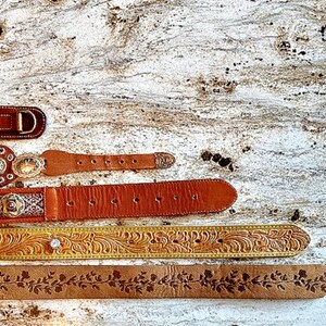 May include: An assortment of leather belts in various styles. The belts are in shades of brown and tan, with decorative buckles and floral or embossed patterns. One belt has a large letter "D" buckle.