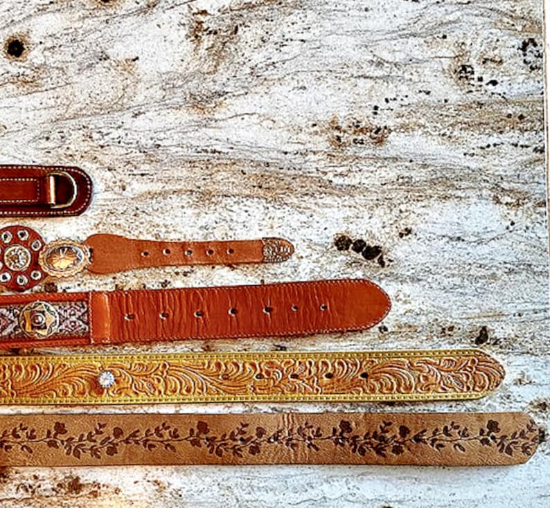 May include: An assortment of leather belts in various styles. The belts are in shades of brown and tan, with decorative buckles and floral or embossed patterns. One belt has a large letter "D" buckle.