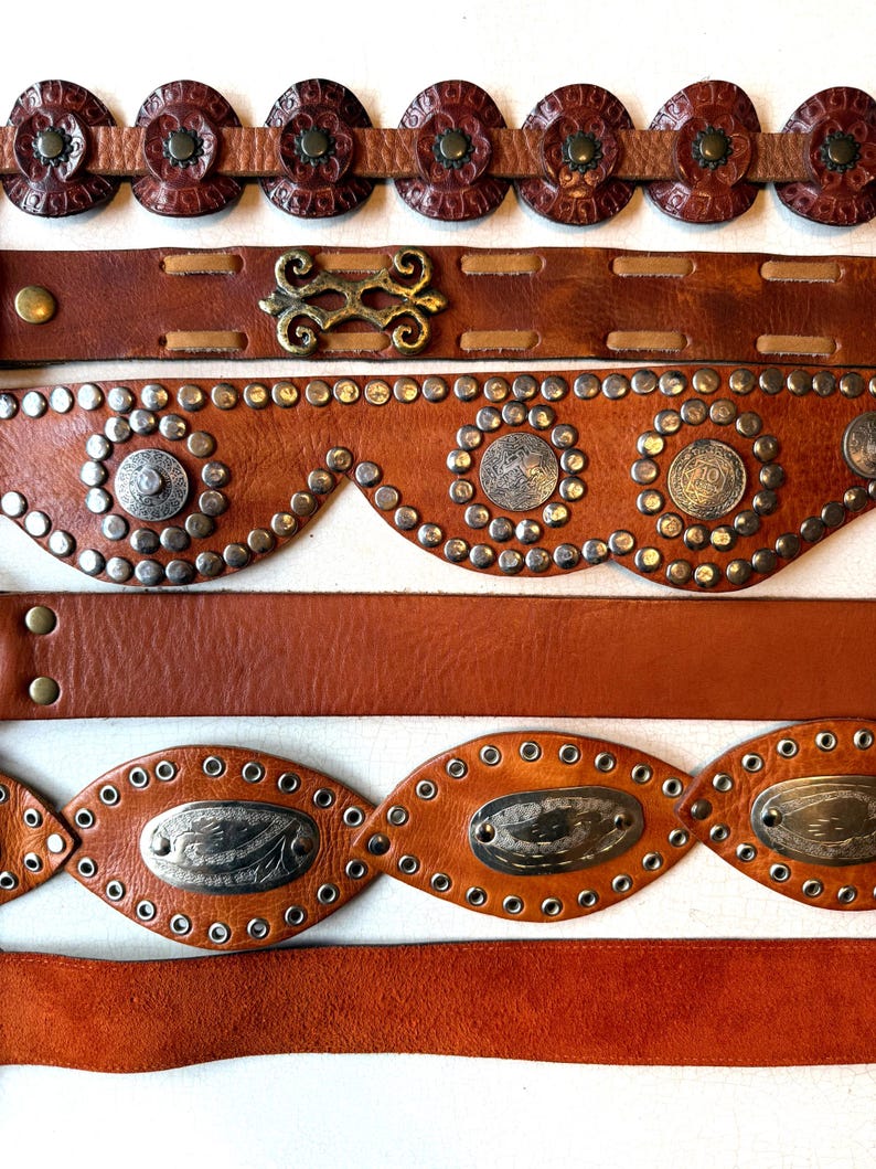 May include: Five brown leather belts with different decorative details. The top belt has round, embossed leather accents. The second belt has a metal buckle with a decorative design. The third belt has a scalloped edge with silver studs and round metal accents. The fourth belt has oval-shaped metal accents with a decorative design and silver studs. The bottom belt is plain.
