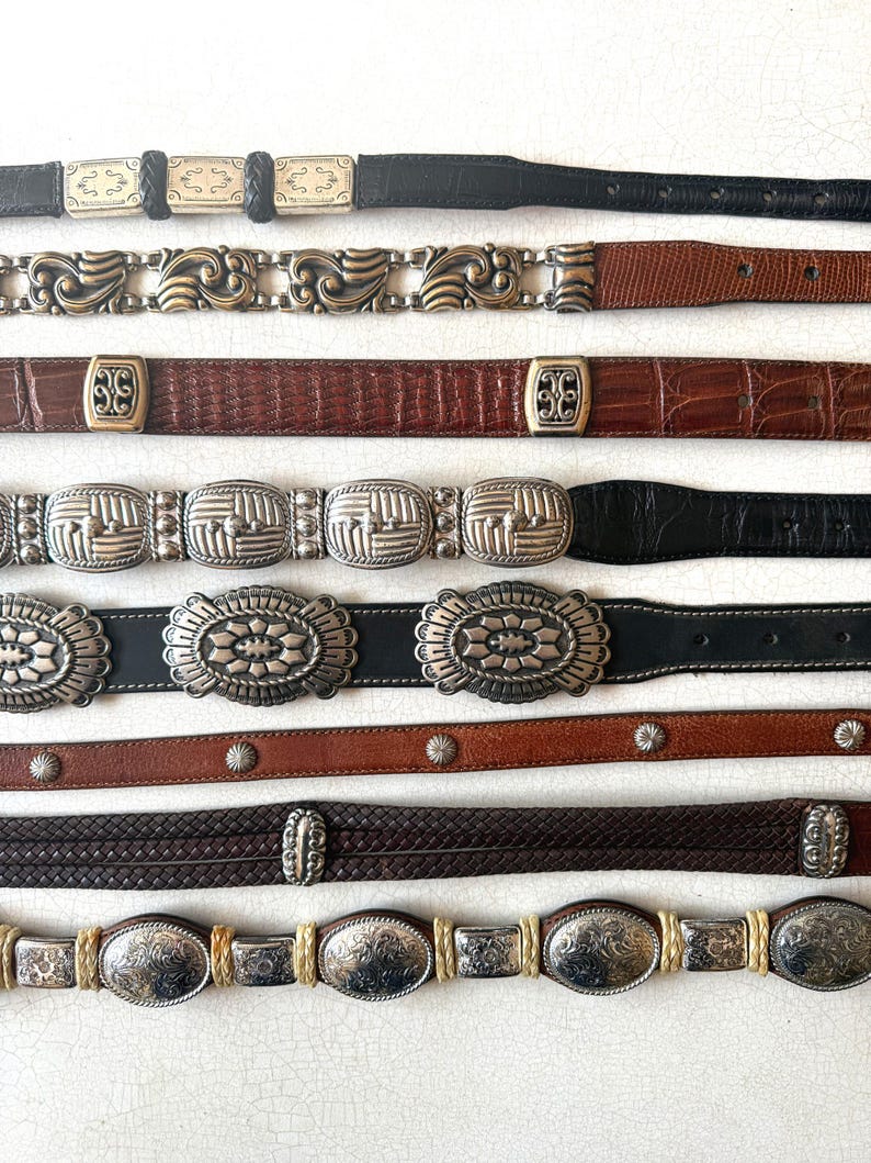 May include: A collection of vintage belts with various designs. The belts feature different colors, including black, brown, and tan. Buckles and decorative elements are silver and gold-toned, with intricate patterns and textures. Styles include braided, embossed, and studded.