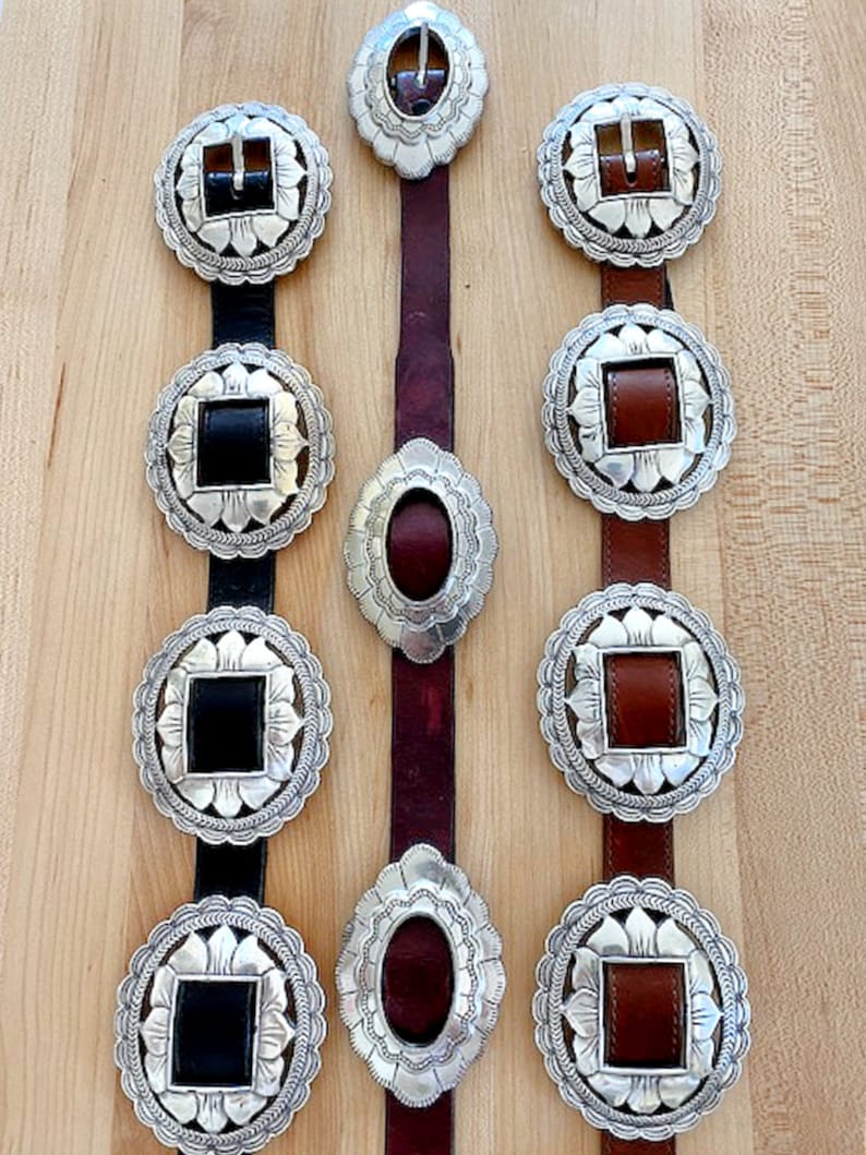 Może przedstawiać: Trzy srebrne klamry do paska z ozdobnymi wzorami kwiatowymi. Klamry są przymocowane do sk&oacute;rzanych pask&oacute;w w kolorze czarnym, brązowym i bordowym.