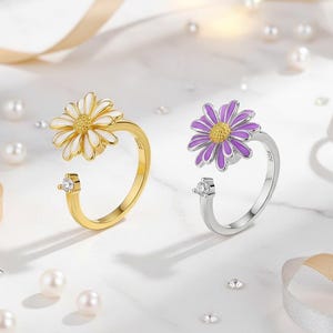 May include: Two daisy flower rings, one gold-toned with a white flower and the other silver-toned with a purple flower. Each ring features a small, clear gemstone. Pearls and ribbons are scattered around the rings.