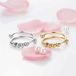 May include: Two open-ended rings, one silver and one gold, each with small, round beads. The rings are set against a backdrop of pink flower petals and small pearls, creating a delicate and elegant aesthetic.