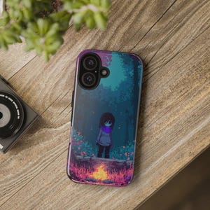 May include: A phone case with a pixelated forest design, showing a character by a campfire. The case has a dark blue, purple, and pink colour scheme. The phone case is on a wooden surface.