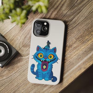 May include: A white phone case featuring a cartoon blue cat with yellow eyes and a bird wearing a hat. The cat has a wide grin and the text "Save the kids" is visible. The case is on a wooden surface.