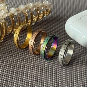 May include: A selection of spinner rings in gold, rose gold, and iridescent finishes. Each ring features a celestial design with moons and stars. The rings are displayed on a textured surface, with a gold hair clip and a white object in the background.
