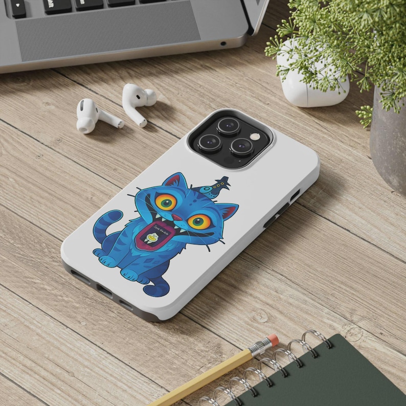 May include: A white phone case featuring a cartoon blue cat with yellow eyes and a party hat. The cat has an open mouth with a graphic inside. The phone case is on a wooden surface with a laptop, earbuds, and a plant in the background.