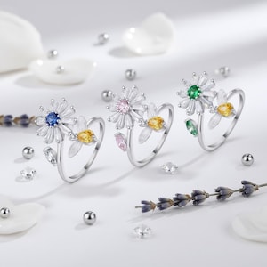 May include: Three silver daisy rings with colourful gemstone centres. Each ring features a different coloured centre stone: blue, pink, and green. Additional yellow and pink gemstones are set on the ring's band, with white petals.
