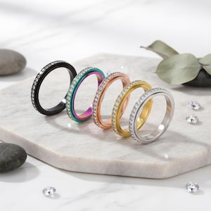 May include: Five rings in various colors: black, rainbow, rose gold, gold, and silver. Each ring is adorned with small, sparkling gemstones. The rings are displayed on a white marble surface, with a few loose gemstones scattered around.