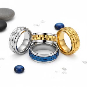 May include: Four metal rings with a textured design. One ring is silver, one is gold, and one is blue. The rings are on a white surface with small blue and grey decorative stones.