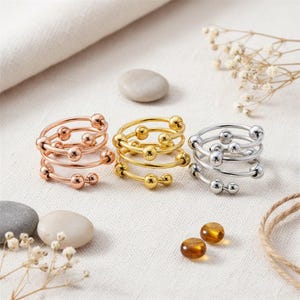 May include: Three spiral rings in rose gold, gold, and silver. Each ring features a coiled design with small, evenly spaced spheres. The rings are displayed on a neutral surface with decorative elements.