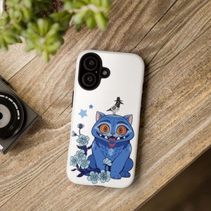 Peut inclure: Coque de téléphone blanche avec un chat bleu de dessin animé aux yeux orange, un oiseau et des motifs floraux bleus. Le chat tire la langue. Deux étoiles bleues sont également présentes.