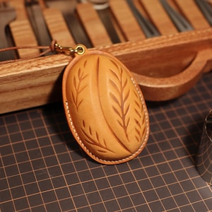 May include: An oval-shaped tan leather case featuring a molded, raised coffee bean design with etched leaf-like patterns on the surface. The case is stitched with white thread along the edges and includes a small brass-colored metal eyelet at the top.