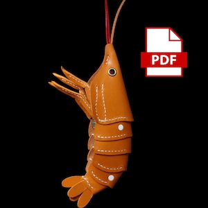 Op de afbeelding: Een handgemaakte, oranje leren garnaal sleutelhanger met witte stiksels en een rode antenne. De garnaal heeft een gesegmenteerd lichaam en gedetailleerde klauwen. Het PDF-pictogram staat rechtsboven.