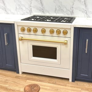 Könnte beinhalten: Miniatur-Herd in Weiß und Gold mit schwarzem Kochfeld, goldenen Knöpfen und einem goldenen Griff. Der Herd wird von marineblauen Schränken flankiert und steht auf einem Holzboden. Eine Münze liegt vor dem Herd auf dem Boden.