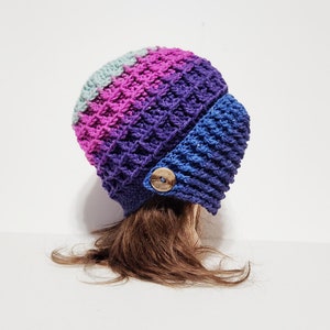 Könnte beinhalten: Eine gehäkelte Mütze mit einem Farbverlauf in Blau, Lila und Rosa. Die Mütze hat eine Klappe, die die Ohren bedeckt, und einen Knopf aus Holz.