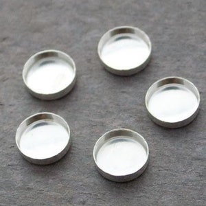 Puede incluir: Cinco pequeñas copas redondas plateadas sobre una superficie gris. Cada copa tiene un interior cóncavo poco profundo, ideal para engarzar piedras o adornos en la fabricación de joyas.