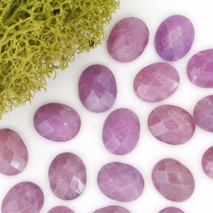 May include: A collection of 18 oval-shaped, faceted, pink gemstones. The stones are arranged in a loose cluster on a white background.