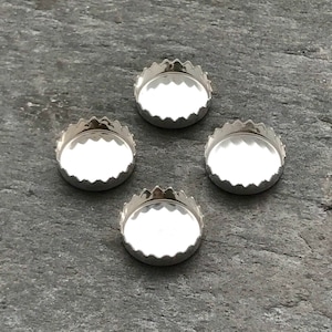 May include: Four silver-colored bezel cups with scalloped edges, arranged on a gray stone surface. The cups have a shallow, concave interior, designed to hold a small object or cabochon. The metal has a bright, reflective finish.