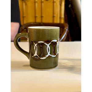 Puede incluir: Taza de cerámica verde oliva con asa curva. La taza presenta un patrón decorativo de círculos blancos entrelazados sobre un fondo marrón. La taza es de tamaño estándar para café o té.