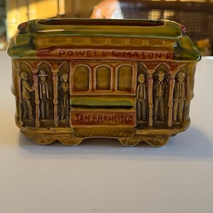 Pode incluir: Uma figura de bonde de cerâmica em tons de castanho, verde e vermelho. As palavras "POWELL & MASON" e "SAN FRANCISCO" são visíveis. Os lados apresentam figuras em relevo de pessoas.
