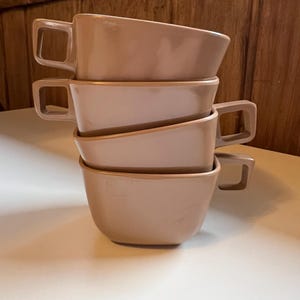 May include: A stack of four beige ceramic mugs with rounded edges and handles. The mugs are stacked on a white surface.