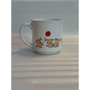 Può includere: Tazza in ceramica bianca con manico curvo. La tazza presenta un giocoso design a cartoni animati di cani che giocano con una palla rossa. I cani sono marroni con contorni neri. La tazza è di dimensioni standard.