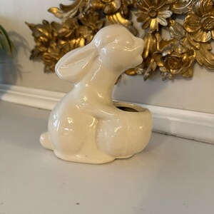Peut inclure: Figurine de lapin en céramique crème avec un centre creux. Le lapin est en position assise, les oreilles dressées. La figurine a une finition brillante et est probablement un objet décoratif.