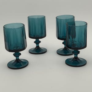 Puede incluir: Cuatro copas de vino de color verde azulado. Cada copa tiene un cuenco cilíndrico con estrías verticales y una base redondeada. El tallo tiene una forma decorativa de reloj de arena. Las copas están dispuestas sobre una superficie blanca.